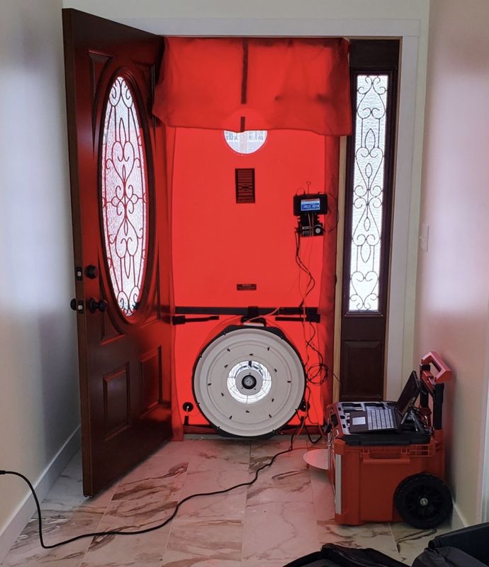 Red blower door test frame and fan set up in a front doorway, with a pressure gauge and laptop on a cart