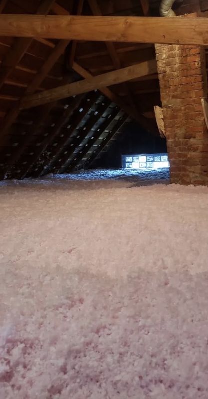 Attic floor covered in pink blown-in fiberglass insulation beside a brick chimney